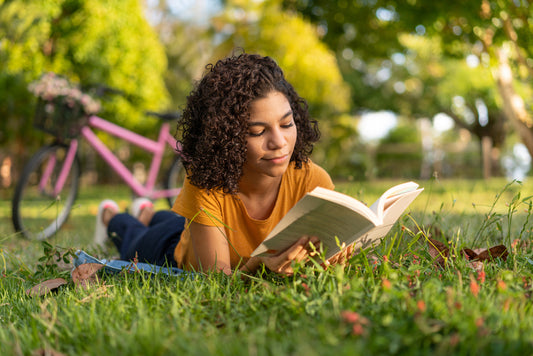 Young adult woman with curly, wavy hair reading a book while lying on the grass. The scene captures a peaceful summer day, ideal for engaging in summer reading