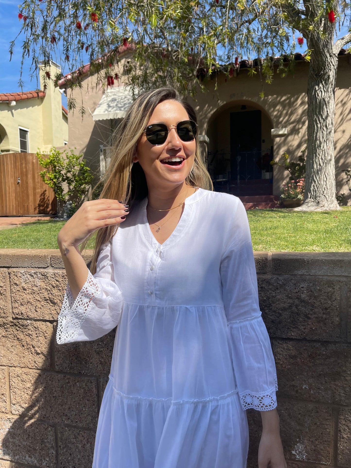 Model smiling outdoors wearing the lightweight boho cotton maxi dress standing in front of a brick building structure, brick wall and grassy area radiating relaxed summer elegance at Tela Designs Store.