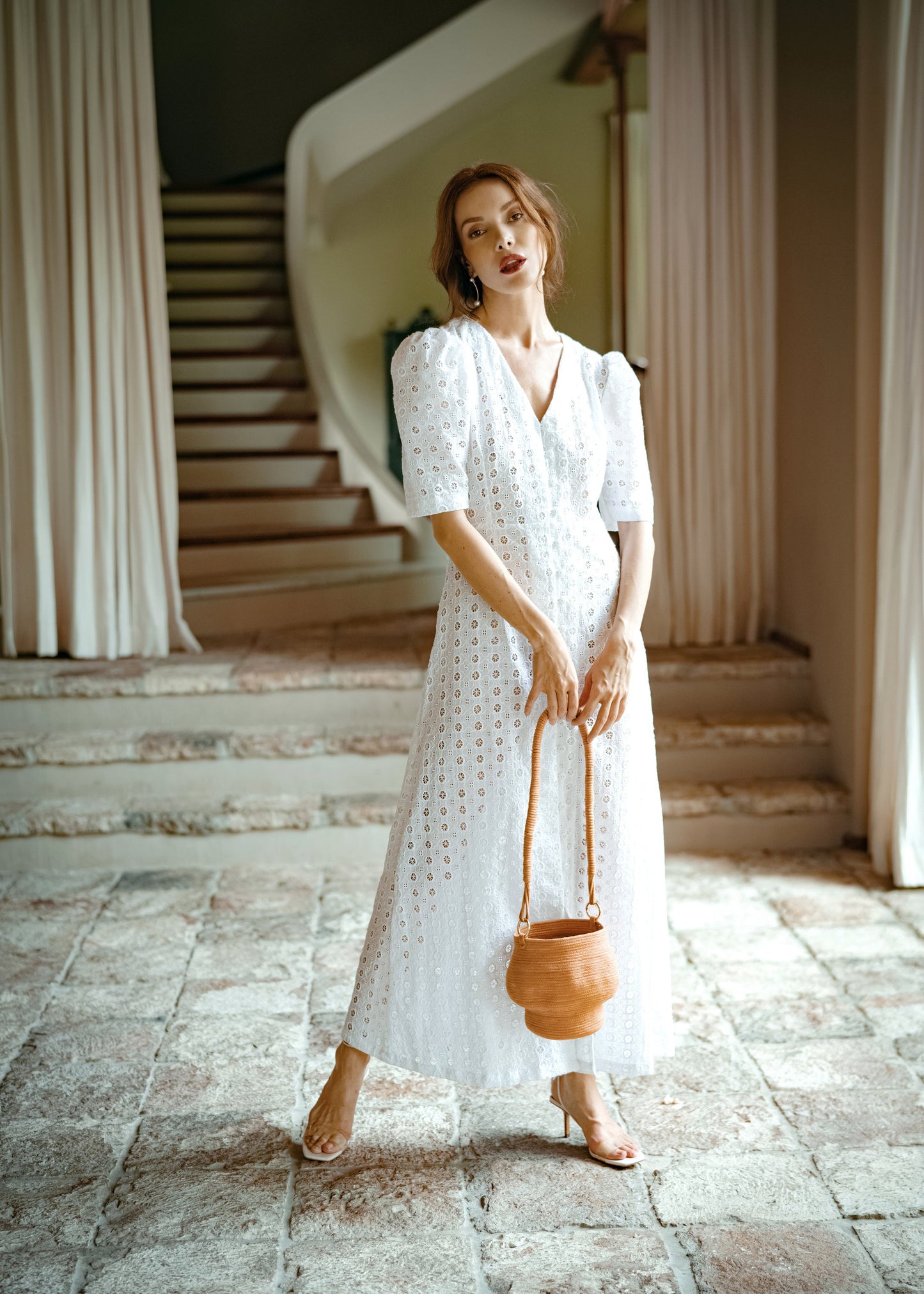 Woman in a white embroidered cotton maxi dress with puff sleeves, holding a brown leather bucket bag in a sunlit stone foyer.