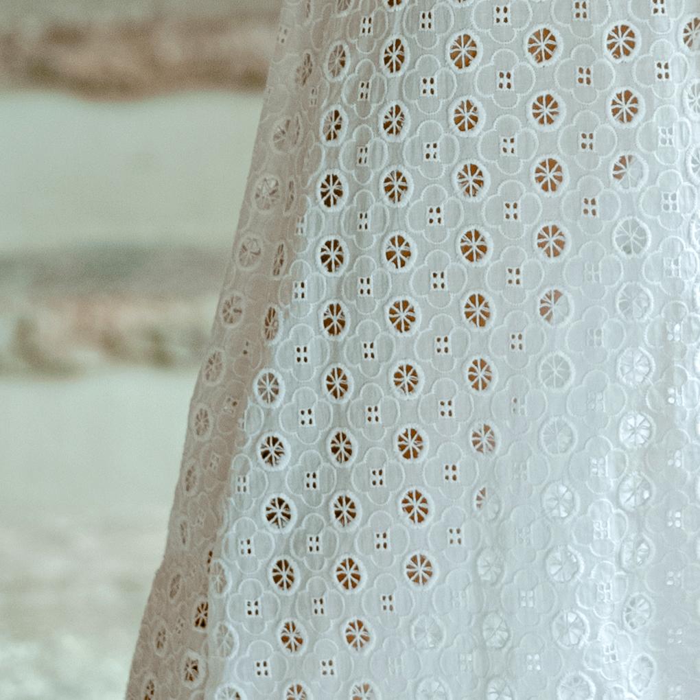 Close-up of white embroidered cotton eyelet fabric with floral clover-like cutouts, showcasing breathable and vintage-inspired texture.