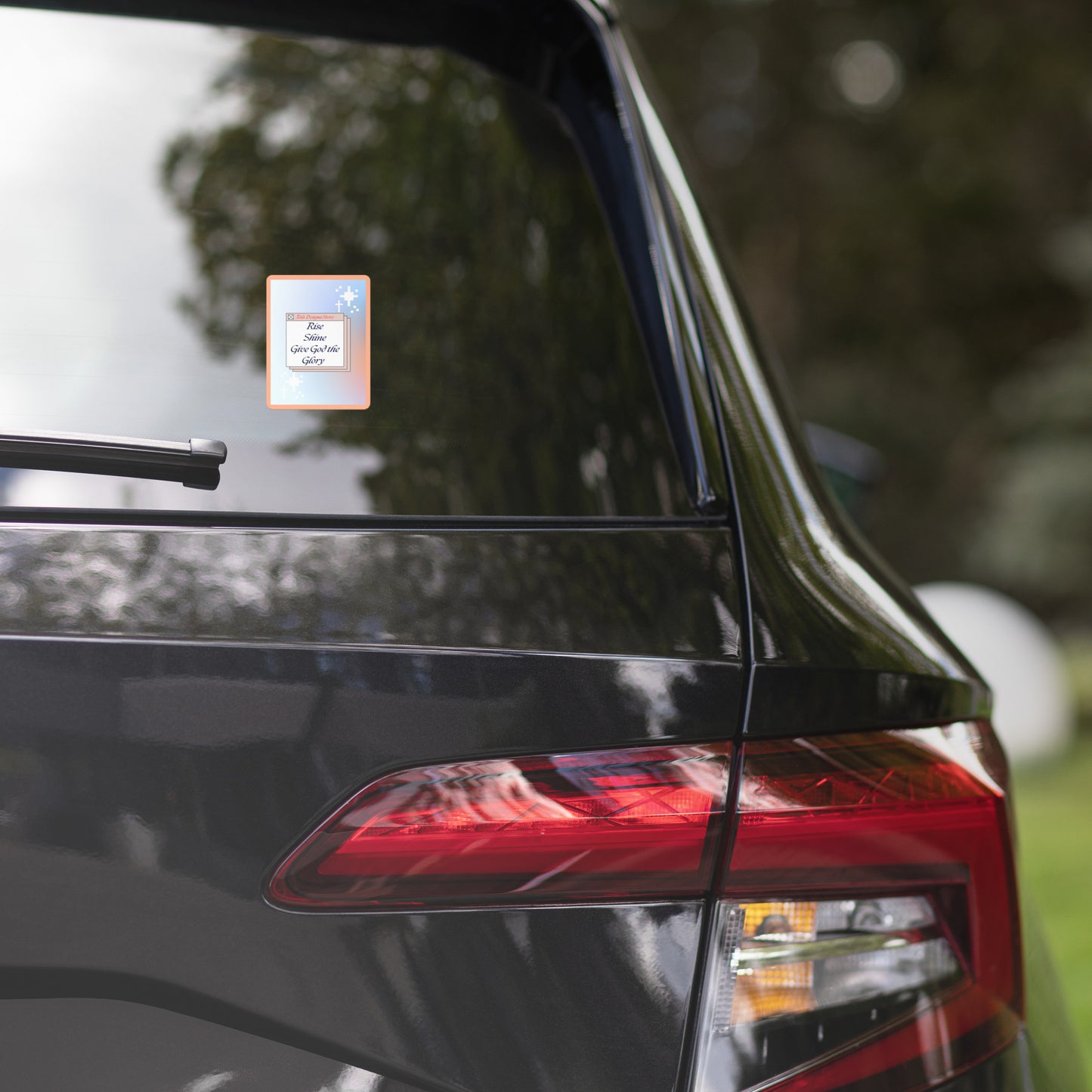 Rise Shine Give God The Glory 15x3.75 holographic sticker with neon orange-pink border and red 'Tela Designs Store' branding displayed on the rear window of a black minivan. The sticker features a bold Christian slogan with a radiant holographic design, perfect for adding a faith-based message to vehicles.