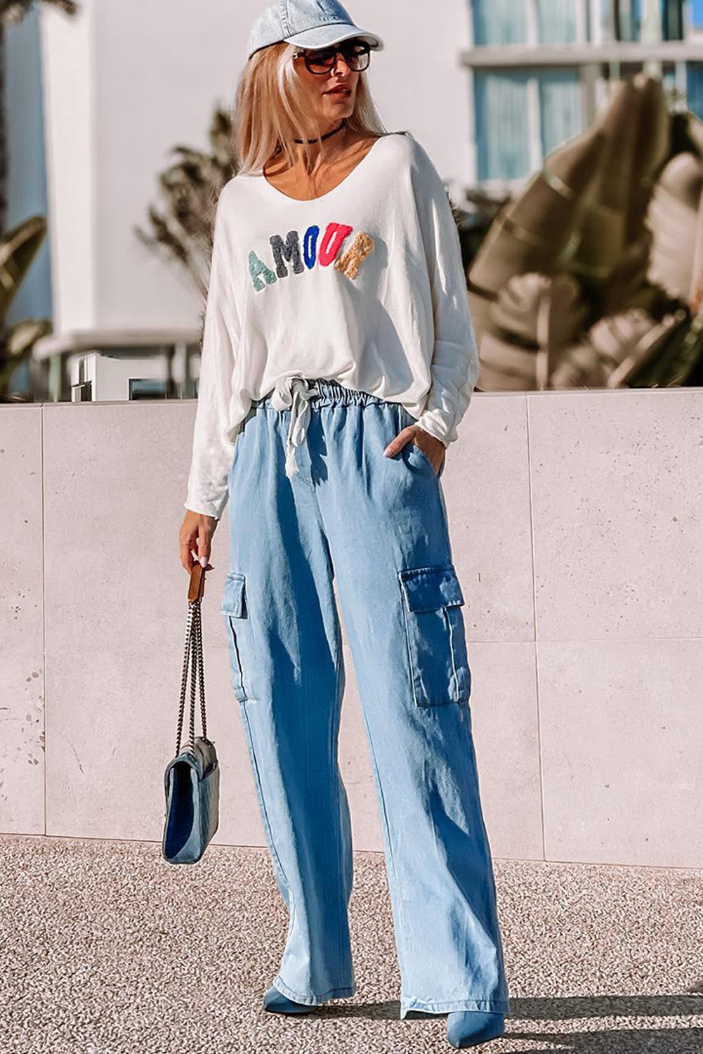 Lifestyle photo of woman in long-sleeve “Amour” graphic top and blue cargo pants styled with heels and matching cap at Tela Designs Store