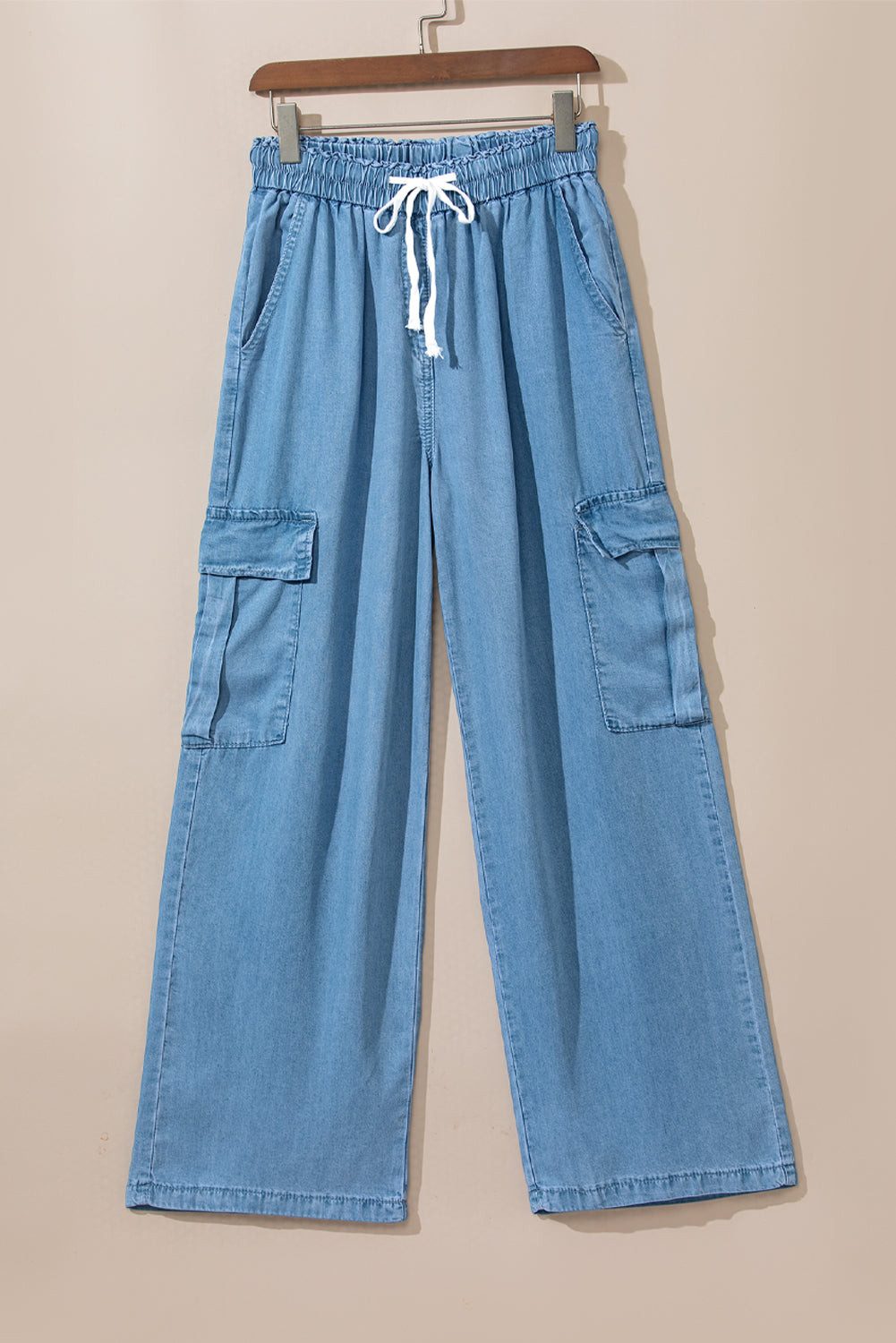 Front view of cotton chambray denim cargo pants with white drawstring and side pockets hanging against neutral wall at Tela Designs Store