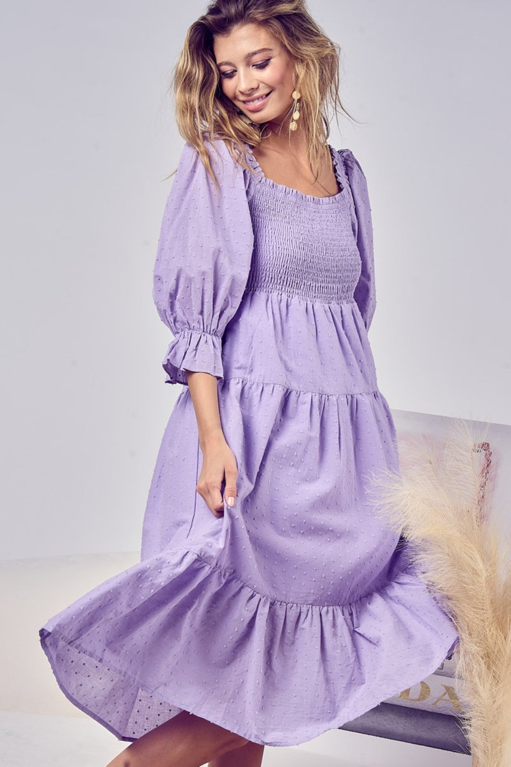Image of a woman wearing the Unapologetic Swiss Dot Smocked Tiered Midi Dress in purple. The model faces sideways to the right, smiling while holding the sides of the dress with her hands. She wears long pearl earrings, and her wavy hair transitions from brown roots to bleached dark blonde ends. She glances downward, and the image shows her from head to just below her knees, with the dress extending slightly beyond.