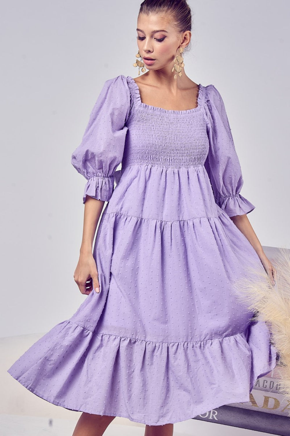 Image of a light skinned black woman wearing the Unapologetic Swiss Dot Smocked Tiered Midi Dress in purple. The model faces front and head to the right, smiling while holding the sides of the dress with her hands. She wears long pearl earrings, and her is pulled back. She glances downward, and the image shows her from head to just below her knees, with the dress extending slightly beyond.