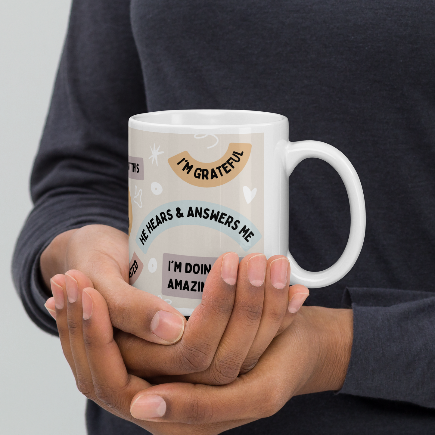 A woman of color shown holding a white ceramic mug in her hands, cupping it that has Christian affirmations printed on it, such as 'I'm grateful' and 'He hears and answers me' (right view). by Tela Designs Store.