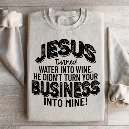 Folded sand-colored Christian fleece sweatshirt with biblical humor text design, "Jesus turned water into wine He didn't turn your business into mine!"
