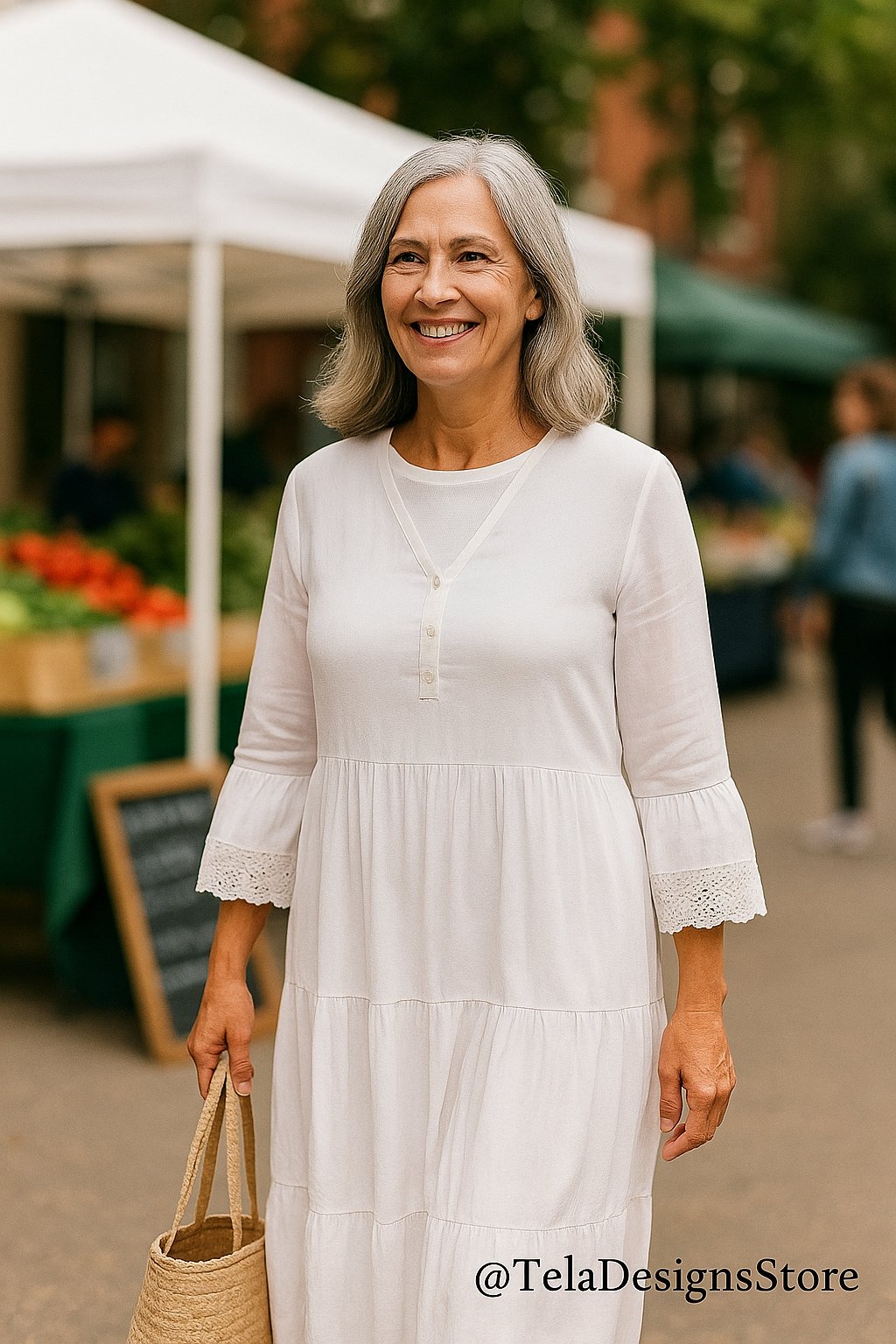 Smiling mature woman wearing a tiered white cotton boho maxi dress with crochet lace sleeves, holding a straw tote while walking through an outdoor market — a joyful, effortless summer look at Tela Designs Store.