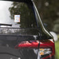 Rise Shine Give God The Glory 15x3.75 holographic sticker with neon orange-pink border and red 'Tela Designs Store' branding displayed on the rear window of a black minivan. The sticker features a bold Christian slogan with a radiant holographic design, perfect for adding a faith-based message to vehicles.