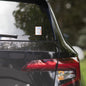 Rise Shine Give God The Glory 3"x3" holographic sticker with neon orange-pink border and red 'Tela Designs Store' branding displayed on the rear window of a black minivan. The sticker features a bold Christian slogan with a radiant holographic design, perfect for adding a faith-based message to car window, water bottles, books, and more!