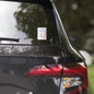 Rise Shine Give God The Glory 4"x4" holographic sticker with neon orange-pink border and red 'Tela Designs Store' branding displayed on the rear window of a black minivan. The sticker features a bold Christian slogan with a radiant holographic design, perfect for adding a faith-based message to vehicles