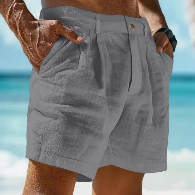 Man wearing gray linen-cotton blend beach shorts with hands in pockets, styled against a blurred beach background. Features button fly.