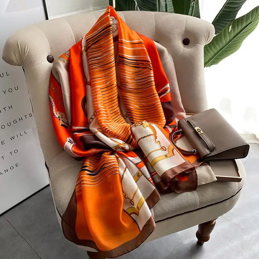 A multicolor humble shawl in the hues of orange, gold, white, and brown is set folded and posed upright in a sitting position like a coat on a gray side sofa chair with a small brown handbag placed neatly on top of it in the seat on the left hand side. There is a tall green potted plant behind the chair with a  bright green leaves noticeable in the bright room with grayish and white tones and a bright patio that is letting in a lot of natural light.