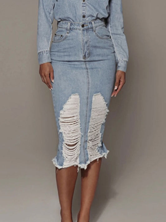Image of a Black female model wearing the Unapologetic Distressed Denim Midi Skirt with Back Slit, paired with a buttoned denim shirt jacket tucked into the skirt. She faces the camera with her hands at her sides, finger nails painted white, and she is shifting her weight onto her right leg. The photo captures her from mid-bust down to her shins, highlighting the midi length of the skirt at Tela Designs Store.