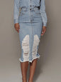 Image of a Black female model wearing the Unapologetic Distressed Denim Midi Skirt with Back Slit, paired with a buttoned denim shirt jacket tucked into the skirt. She faces the camera with her hands at her sides, finger nails painted white, and she is shifting her weight onto her right leg. The photo captures her from mid-bust down to her shins, highlighting the midi length of the skirt at Tela Designs Store.