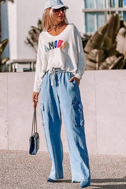 Lifestyle photo of woman in long-sleeve “Amour” graphic top and blue cargo pants styled with heels and matching cap at Tela Designs Store