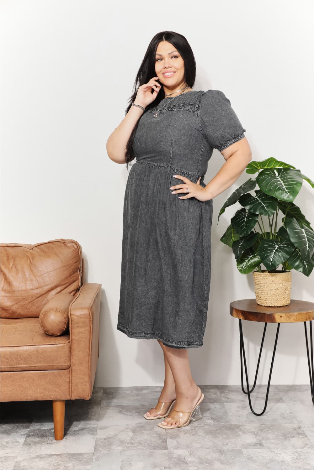 Image of a full-figured woman wearing the Unapologetic Chambray Elegance Midi Dress in gray. She stands smiling in a room between a couch and a table with a potted plant. Facing the camera with her left side, she touches her right ear with her right hand and places her left hand on her hip. Her left toe points down toward the floor. She wears skin-toned low heels.