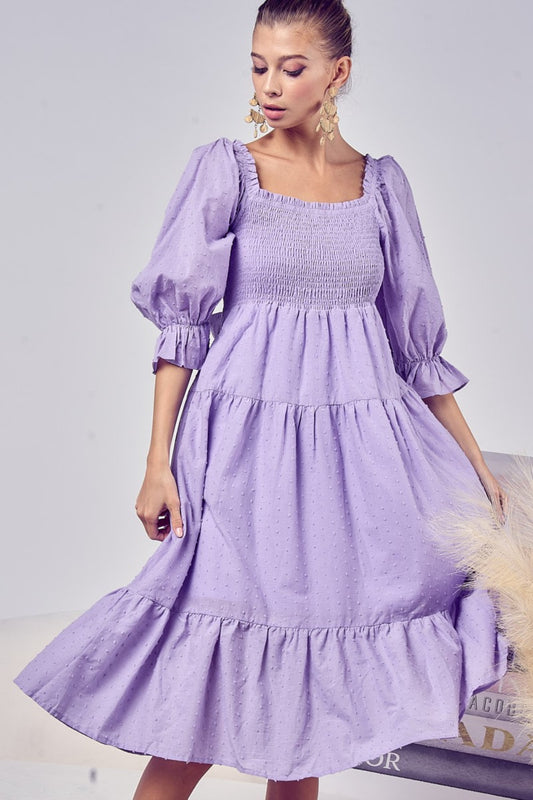 Image of a light skinned black woman wearing the Unapologetic Swiss Dot Smocked Tiered Midi Dress in purple. The model faces front and head to the right, smiling while holding the sides of the dress with her hands. She wears long pearl earrings, and her is pulled back. She glances downward, and the image shows her from head to just below her knees, with the dress extending slightly beyond.